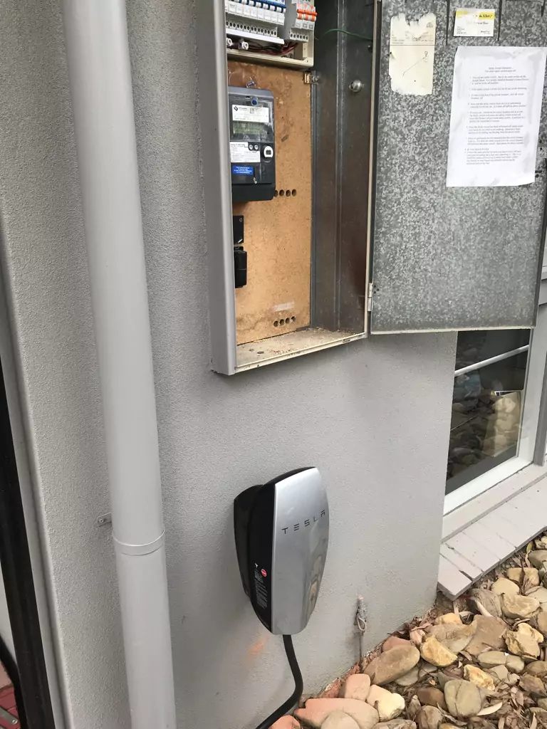 Tesla wall connector installed next to an open electrical panel on an exterior wall.